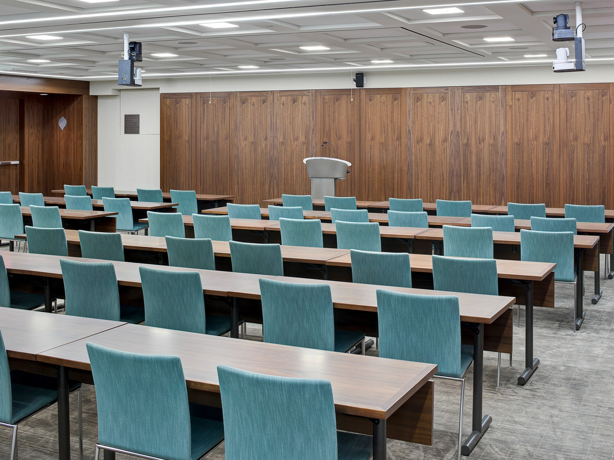 Modern conference room featuring rows of turquoise chairs, dark wood-paneled walls, and a sleek lecturer's podium. Overhead, linear lighting complements the room's clean lines.