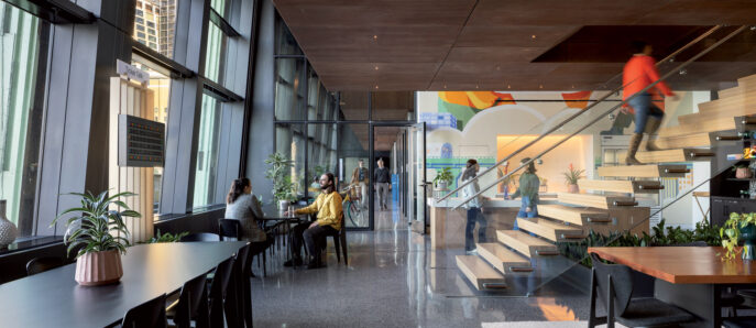 Modern office lobby with polished concrete floors, wood-paneled walls, and full-height windows. Sleek, black communal tables pair with minimalist chairs. A glass-enclosed staircase adds an airy feel, while indoor plants introduce a touch of greenery.
