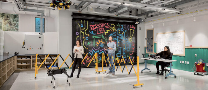 Modern robotics lab interior with exposed ceiling conduits, vibrant graffiti art wall, two standing individuals interacting with a quadruped robot, and a seated person at a desk in the background. Industrial-style workspace with creative, collaborative atmosphere.