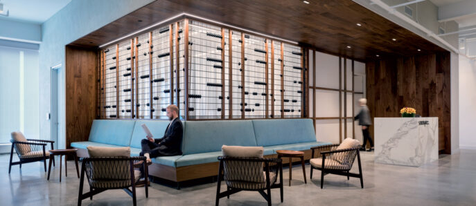 Spacious lobby with polished concrete floors, featuring an expansive wooden wall with built-in shelves displaying black linear elements. Central teal upholstered bench seating is flanked by mid-century modern chairs, anchored by a marble welcome desk, under a high vaulted ceiling with exposed beams.