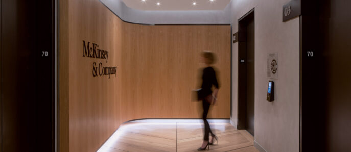 A sleek, modern office corridor featuring warm wooden panels and recessed ceiling lights; signage for 'McKinsey & Company' adorns a door, while a motion blur of a person adds dynamism to the composed setting.