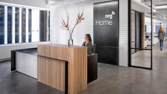 Modern, minimalist reception area featuring a sleek, wooden desk with a clear vase of tall branches. The design incorporates ample natural light, clean lines, and a neutral color palette, complemented by industrial-style doors and concrete flooring.