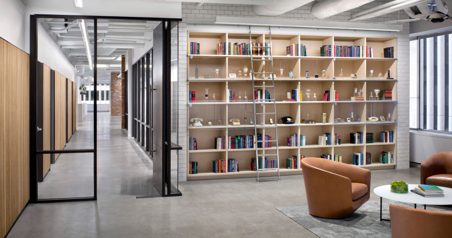 Open-plan office featuring a neutral color palette with natural wood accents, black-framed glass partitions, and an extensive wall-mounted bookshelf. Mid-century modern seating and industrial-style lighting complement the contemporary feel of the space.