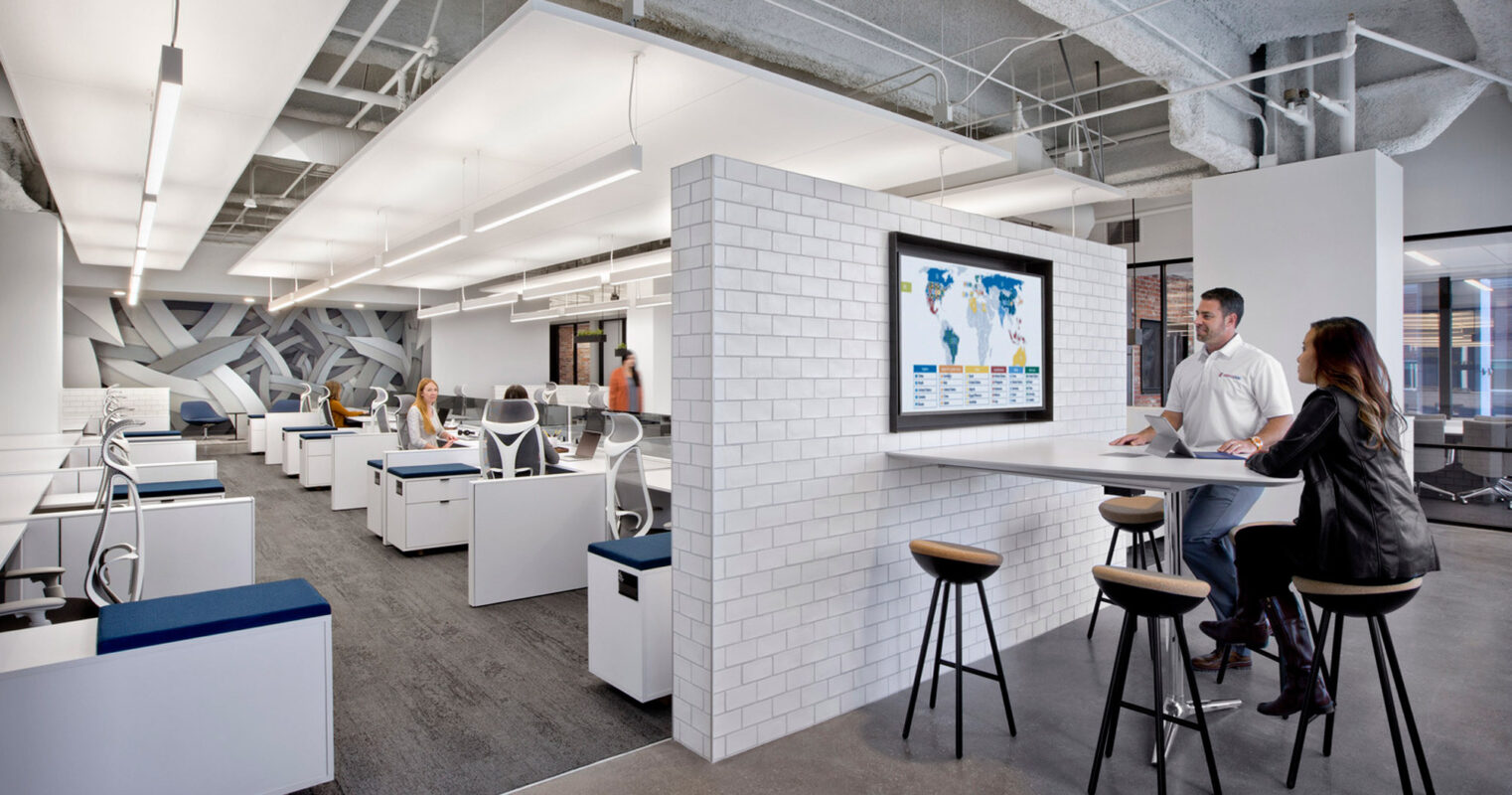Contemporary office space with exposed ceiling showcasing industrial ductwork. The central white tiling feature wall contrasts with the raw concrete, while ergonomic furnishings and sophisticated lighting contribute to a dynamic work environment.