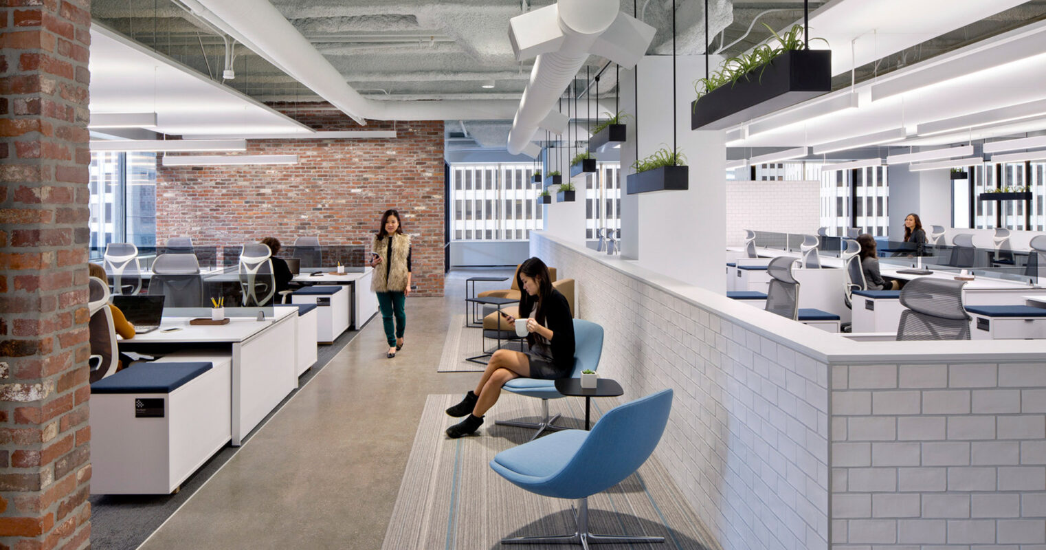 Modern open-plan office with exposed ceiling showcasing ductwork, track lighting, and acoustical baffles. Brick accent walls blend with sleek white workstations and pops of blue in seating. Occupants navigate the space, underscoring the functional design.