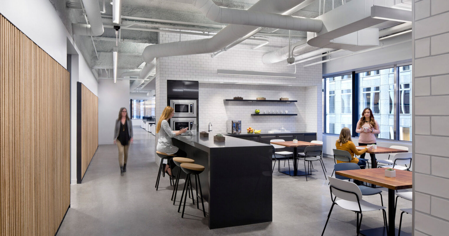 Modern office break room featuring an open-plan layout with polished concrete floors, wooden accents, and a central kitchen island. Exposed ceiling pipes and ductwork contribute to an industrial aesthetic, complemented by natural light from large windows. Employees casually interact in the multifunctional, contemporary workspace.