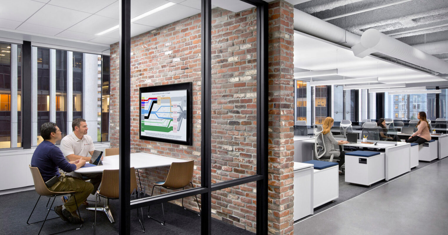 Modern open-plan office with exposed brick walls, incorporating a balance of natural and industrial design elements. Individuals collaborate at a communal table, while others work at standing desks against a backdrop of floor-to-ceiling windows with urban views.