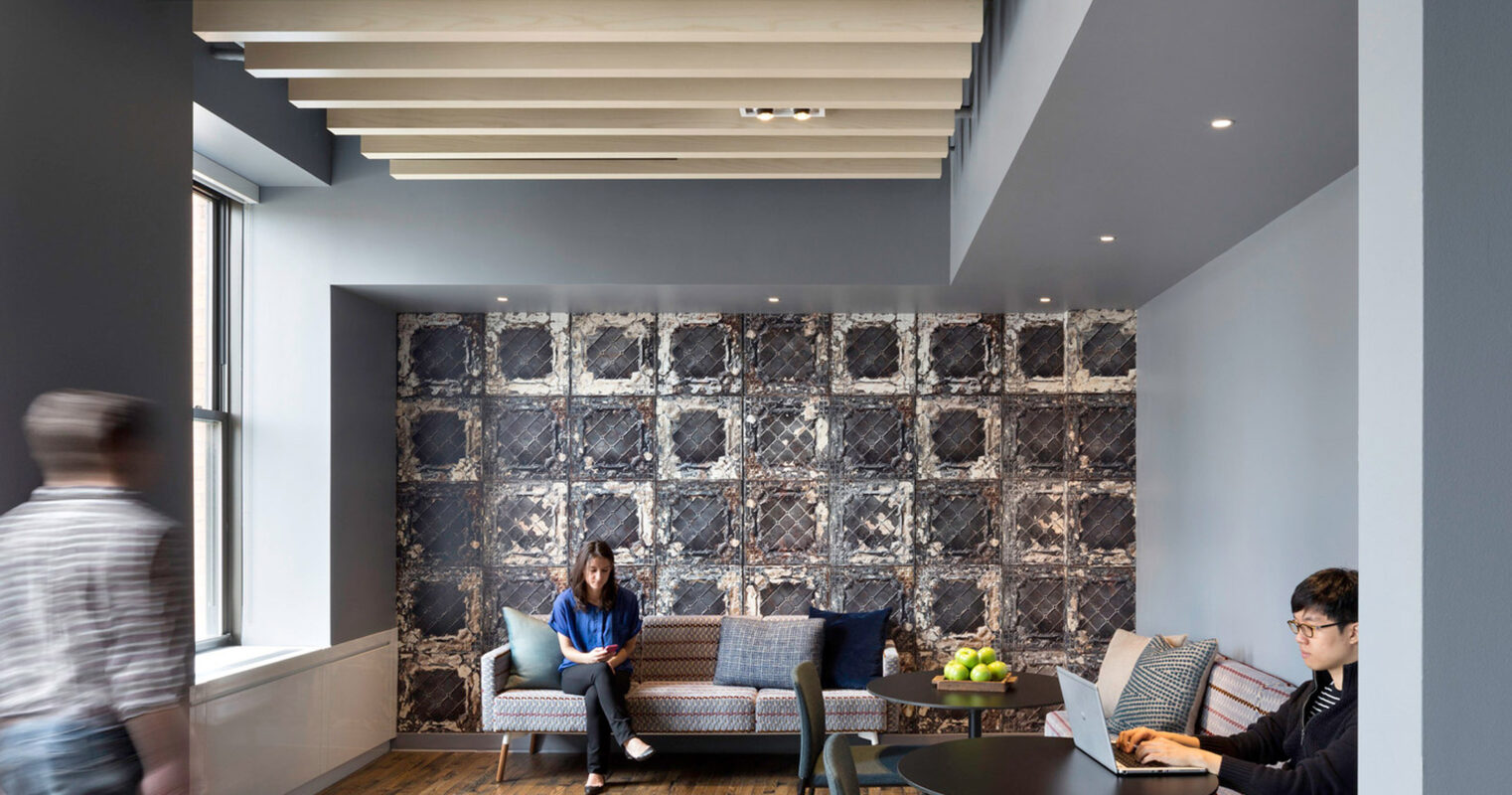Open-plan office space featuring a geometric patterned accent wall, suspended linear lighting, and contrasting deep blue and neutral tones. Seating includes a textured gray sofa and armchairs, complementing the industrial-style exposed ceiling beams. Occupants engage in individual tasks, emphasizing the room's functionality.