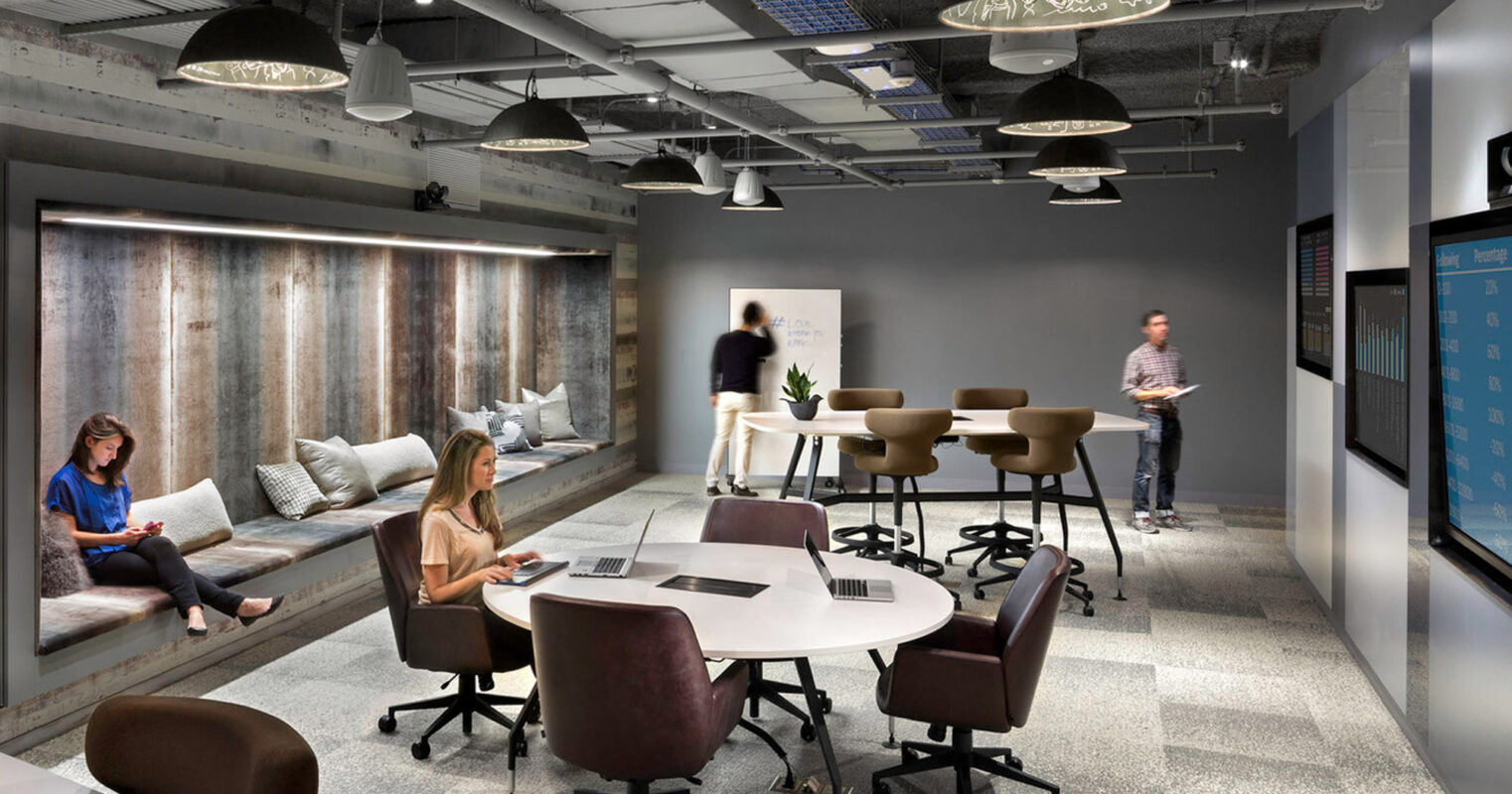 Modern office space blends industrial and rustic elements, featuring exposed ceilings, pendant lighting, distressed wood accents, and ergonomic furniture for a collaborative environment. People work comfortably at communal tables and lounge areas.
