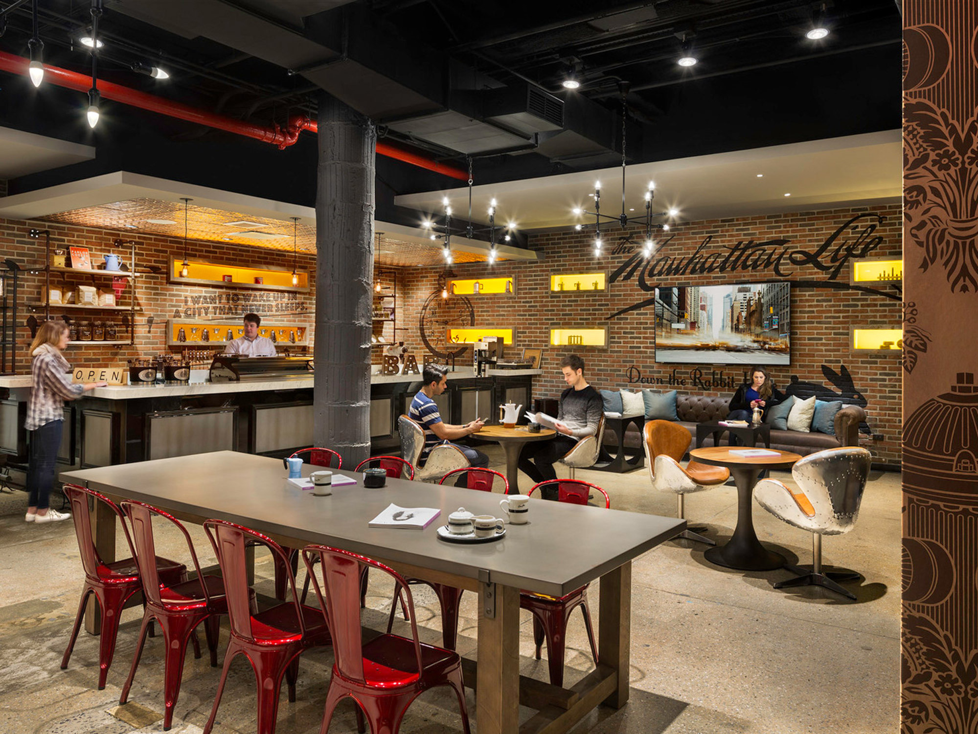 Modern eclectic cafe interior with industrial exposed ceiling and red accents. Mix of communal and private seating options, highlighted by warm pendant lighting above a polished concrete floor. Graphic wall treatments add a vibrant contrast to the urban aesthetic.
