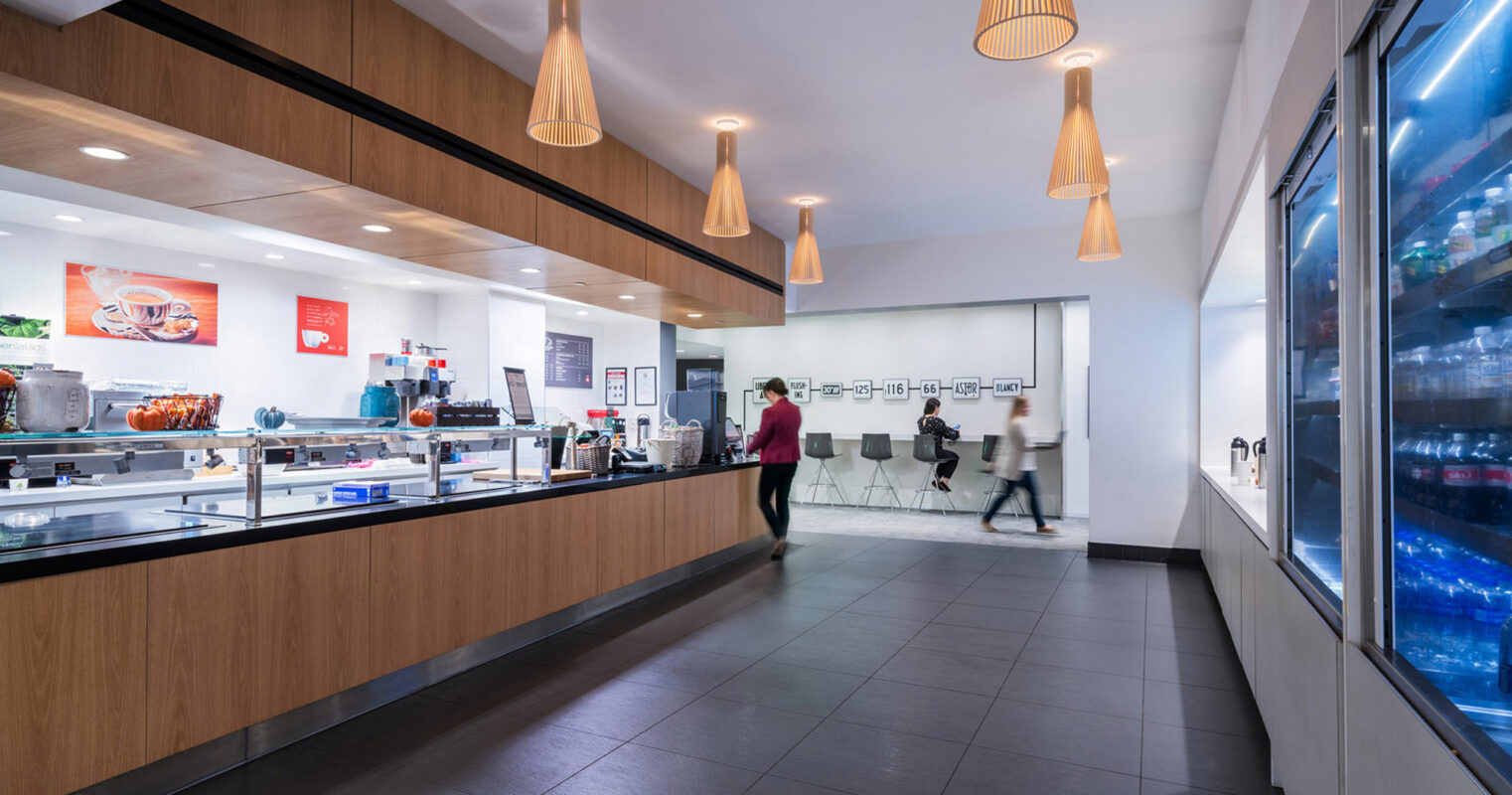 Modern cafeteria featuring a sleek white countertop, wooden accents, and stylish pendant lighting, with a beverage cooler on one side and seating area in the backdrop.
