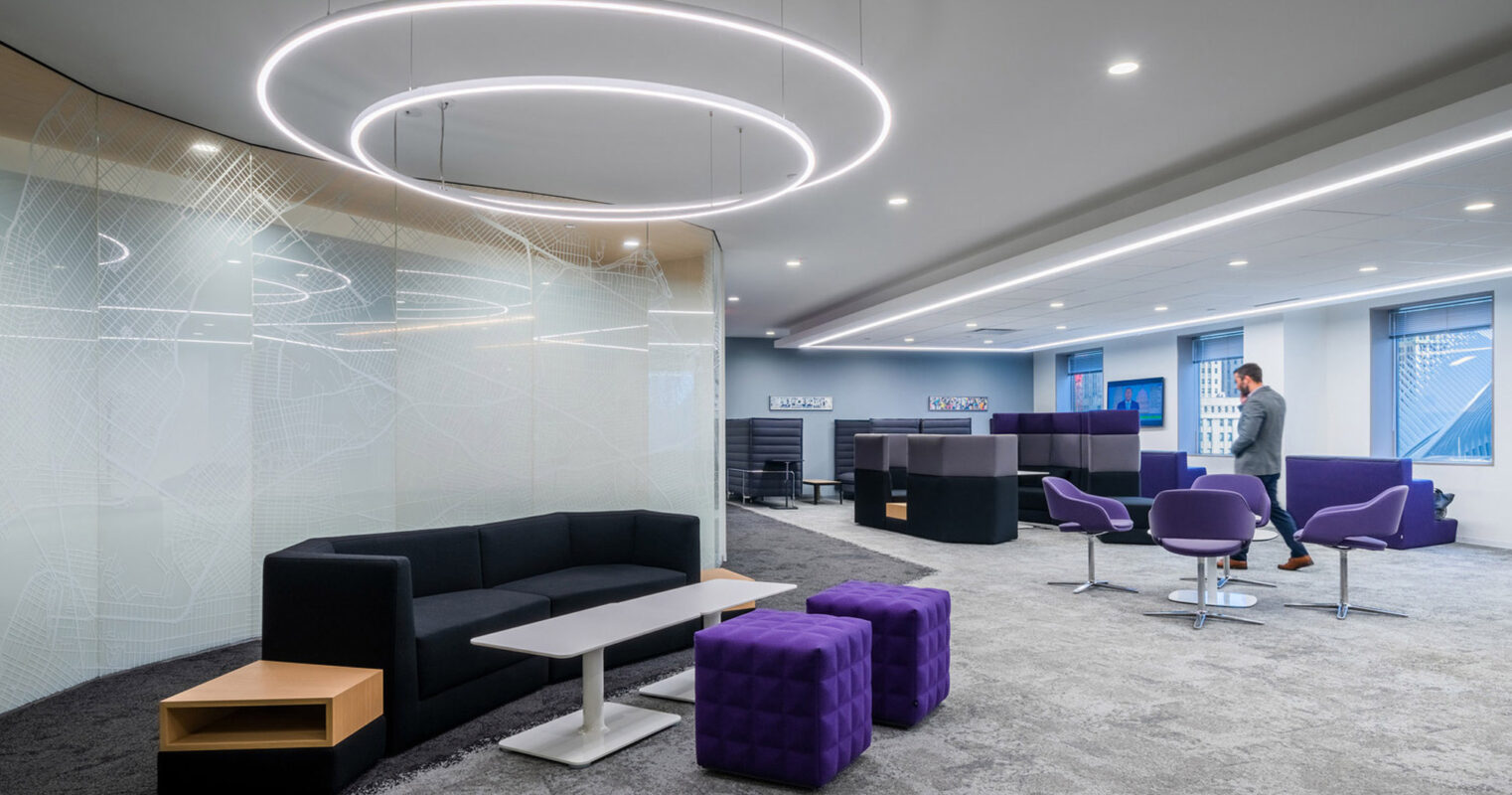Modern office lounge featuring a sleek black sofa, plum-colored armchairs, and vibrant purple ottomans beneath an oversized circular light fixture. The setting is accentuated by geometric patterns adorning glass partitions, with a gentleman in business attire observing the city view.