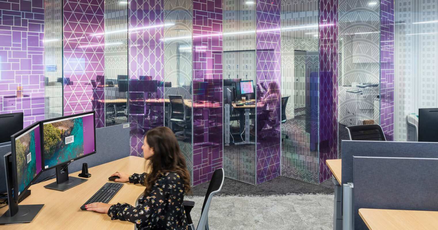 Modern office interior featuring ergonomic workstations with dual monitors, accented by geometric-patterned glass partitions in purple tones, providing both privacy and a visually stimulating environment for productivity.
